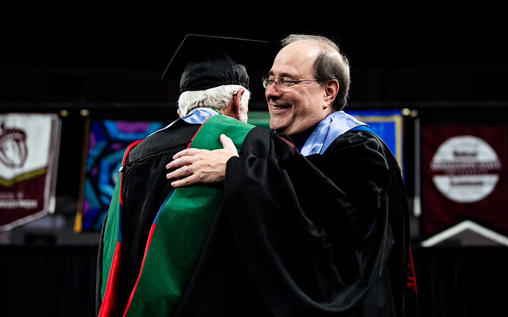 Two people in academic regalia smile and embrace at a graduation ceremony, with colorful university banners visible in the background.