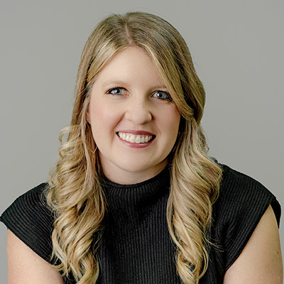 Woman with long blonde curly hair wearing a black short-sleeve top, smiling at the camera against a plain gray background.