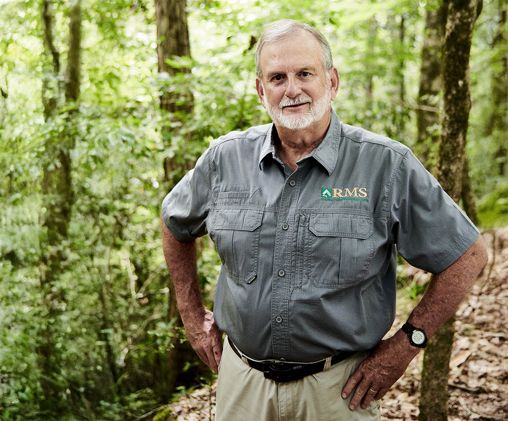 An older man with gray hair and beard stands in a forest, wearing a gray short-sleeve button-up shirt with an "RMS" logo, khaki pants, and a watch.