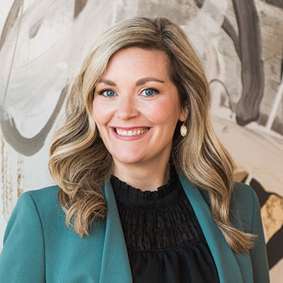 A woman with long blonde hair wearing a teal blazer and black top smiles at the camera in front of an abstract background.