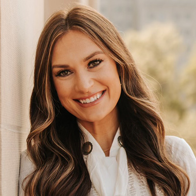 A woman with long brown hair and a white blouse smiles while standing outdoors next to a light-colored wall.