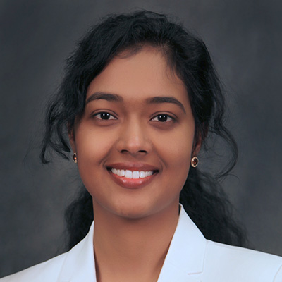A woman with long dark hair, wearing a white blazer and small gold earrings, smiles at the camera against a dark gray background.