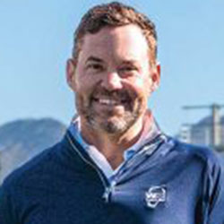 A man with short brown hair and a beard, wearing a blue quarter-zip sweater, stands outdoors with mountains and a clear sky in the background.