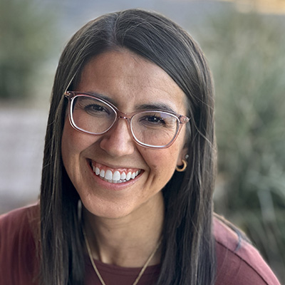 Woman with long dark hair and glasses smiles at the camera while standing outdoors in natural light.