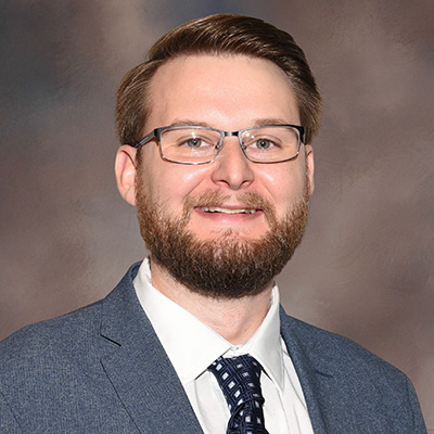 A man with short brown hair, a beard, and glasses is wearing a blue suit, white shirt, and patterned tie, smiling in front of a cloudy gray background.
