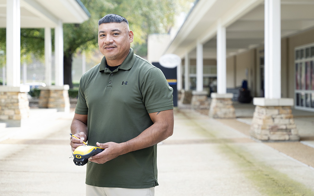 A man in a green polo shirt stands outdoors holding a small yellow and black device, with buildings and columns in the background.