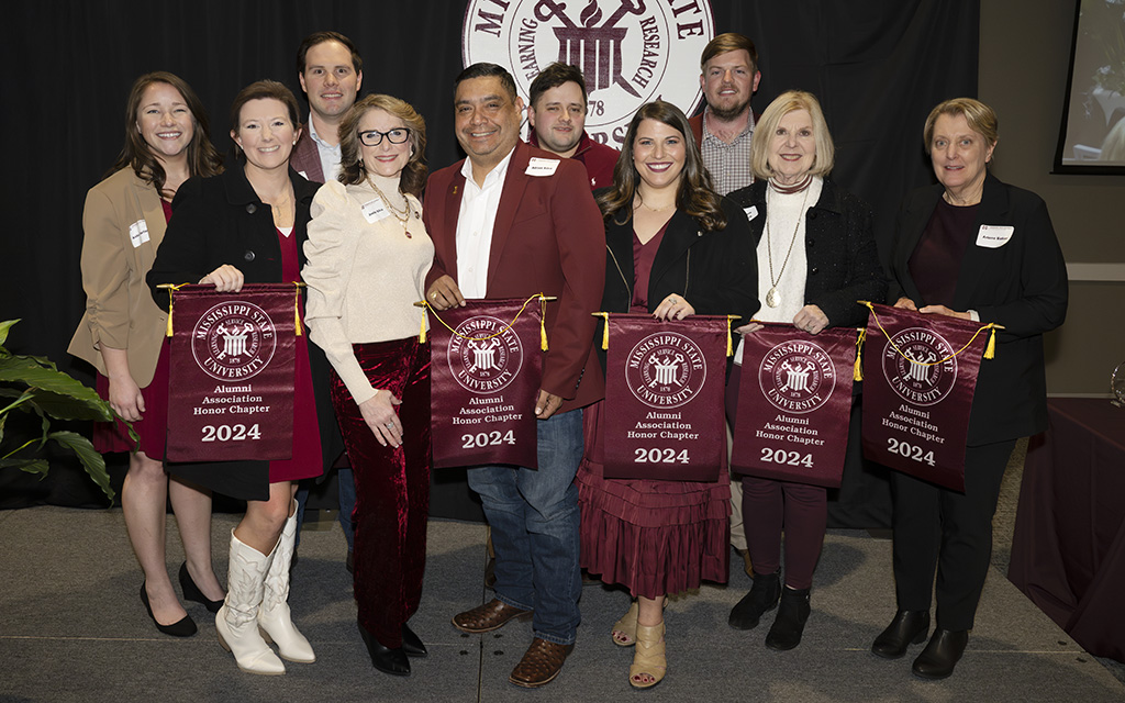 A group of people stand indoors, holding maroon banners that read "Alumni Association Honor Chapter 2024" with the Mississippi State University seal.