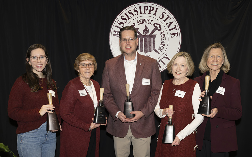 Five people stand in front of a Mississippi State University sign, each holding a cowbell award and wearing name tags and maroon attire.