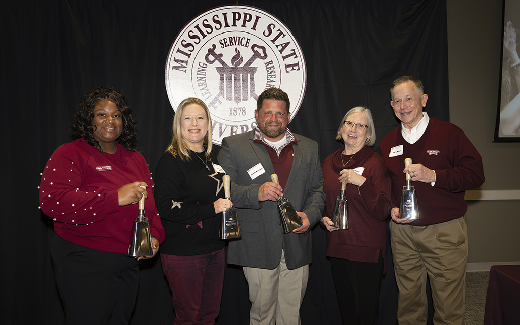 Five adults stand in front of a Mississippi State University sign, each holding an award and smiling at the camera.