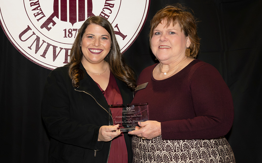 Two women stand together in front of a university emblem. One is holding a glass award plaque and both are smiling at the camera.