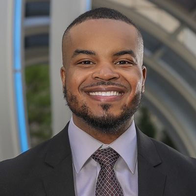 A man in a suit and tie smiles at the camera, standing outdoors in front of a modern architectural structure.