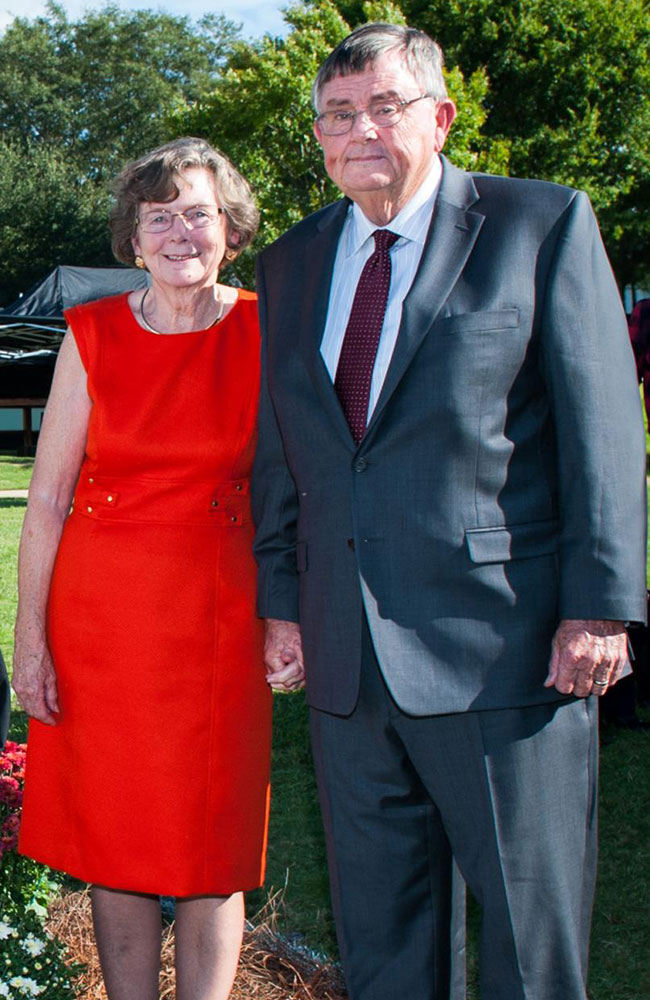 An older woman in a sleeveless red dress stands next to an older man in a dark suit and tie outdoors, both smiling at the camera.