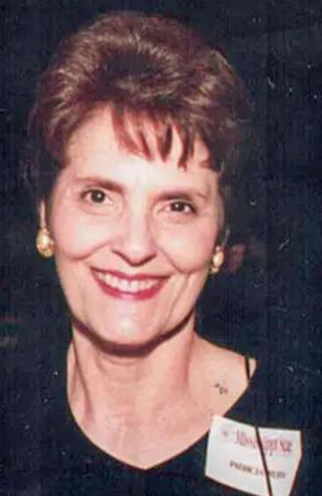 A woman with short brown hair and gold earrings smiles at the camera, wearing a dark top and a name tag.
