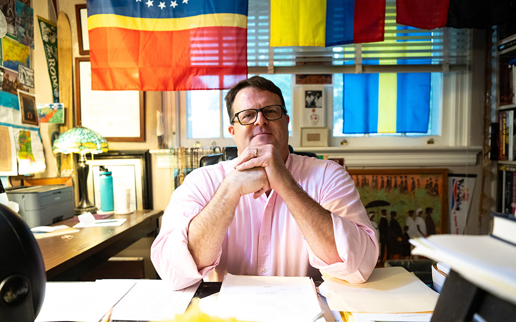 A man in a pink shirt sits at a desk with papers, surrounded by books, maps, and several colorful flags hanging behind him.