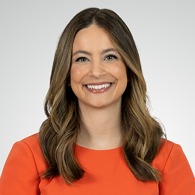 Woman with long brown hair wearing an orange top, smiling at the camera against a plain light gray background.