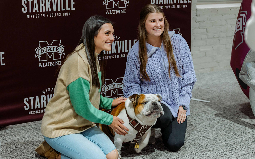 Two women kneel next to a bulldog mascot wearing a maroon vest in front of a Mississippi State University alumni backdrop.