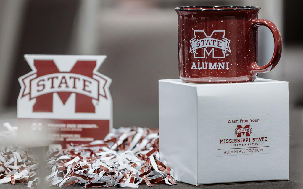 Maroon Mississippi State University Alumni mug on a white box, with MSU logo sign and maroon and white shredded paper in the background.