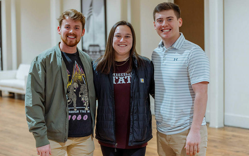Three young adults stand indoors, smiling at the camera. The person in the center wears a "Mississippi State" shirt; the others wear casual jackets and shirts.