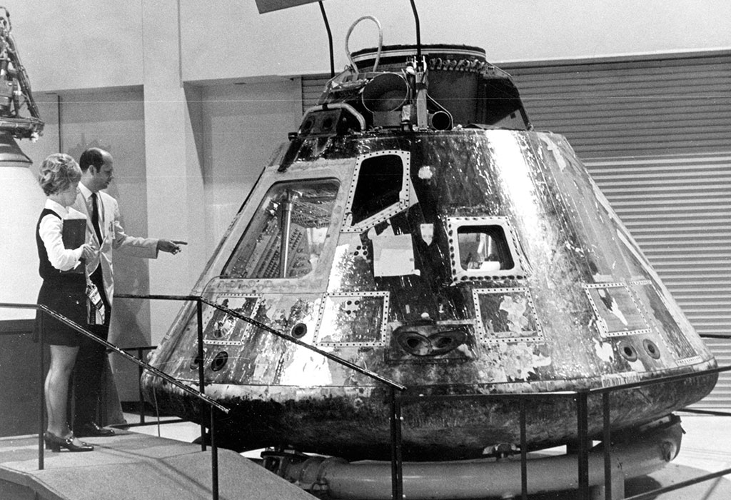 Two people stand next to a NASA Apollo command module on display indoors, examining its exterior through protective railings.