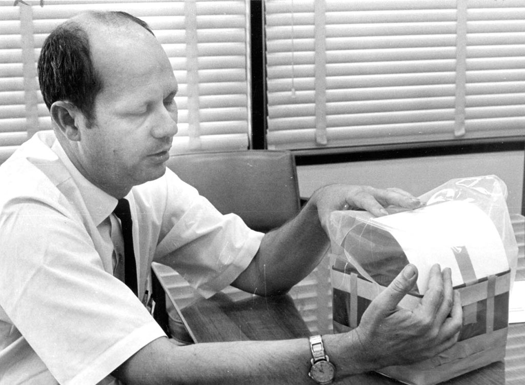 A man in a dress shirt and tie sits at a table, examining a box wrapped in plastic with documents on top, in front of window blinds.