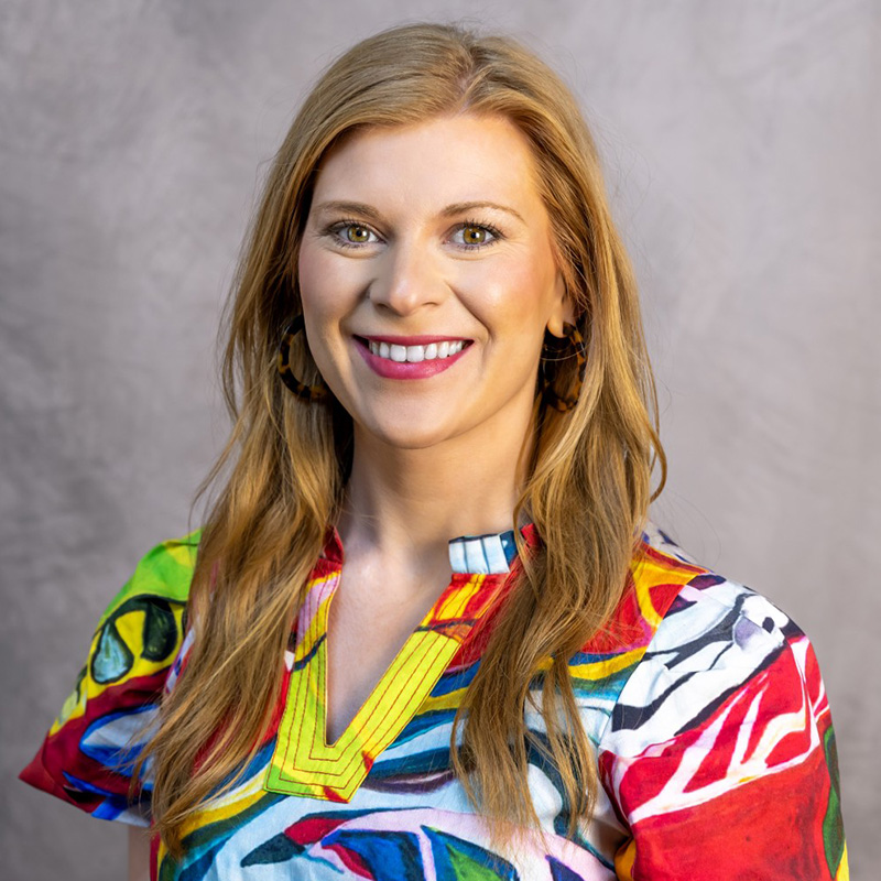 A woman with long light brown hair smiles at the camera, wearing a colorful, patterned blouse and hoop earrings, against a neutral, textured background.