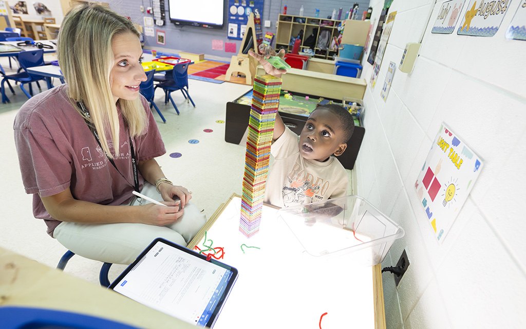 A young child stacks colorful blocks into a tall tower on a light table while a teacher observes and takes notes in a classroom setting.