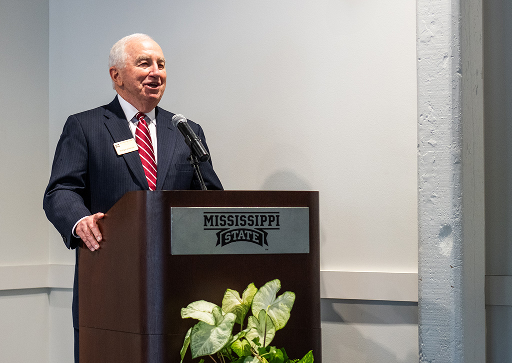 An older man in a suit speaks at a podium with a "Mississippi State" sign; a potted plant is in front of the podium.