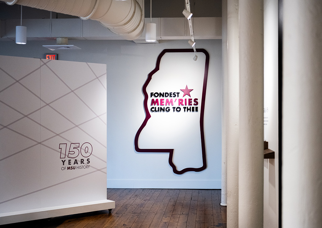Exhibit space with a wall display featuring the outline of Mississippi and text reading “FONDEST MEMORIES CLING TO THEE,” alongside a sign marking 150 years of MSU history.