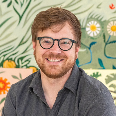 A smiling man with glasses and a beard sits in front of a colorful, floral-patterned background.