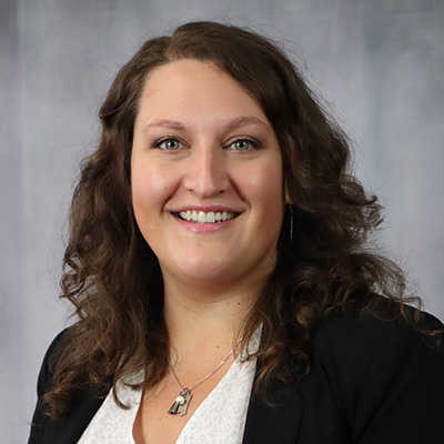 A woman with wavy brown hair wearing a black blazer and white blouse smiles at the camera in front of a gray background.