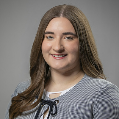 A woman with long brown hair, wearing a gray jacket over a white top, poses in front of a plain gray background.