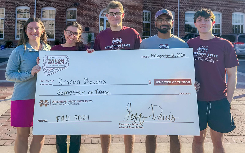 Five people stand outside holding a large check for a semester of tuition awarded to Brycen Stevens from Mississippi State University Alumni Association, dated November 8, 2024.