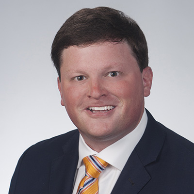 Man with short brown hair wearing a dark suit jacket, white shirt, and yellow striped tie, smiling at the camera against a plain light background.