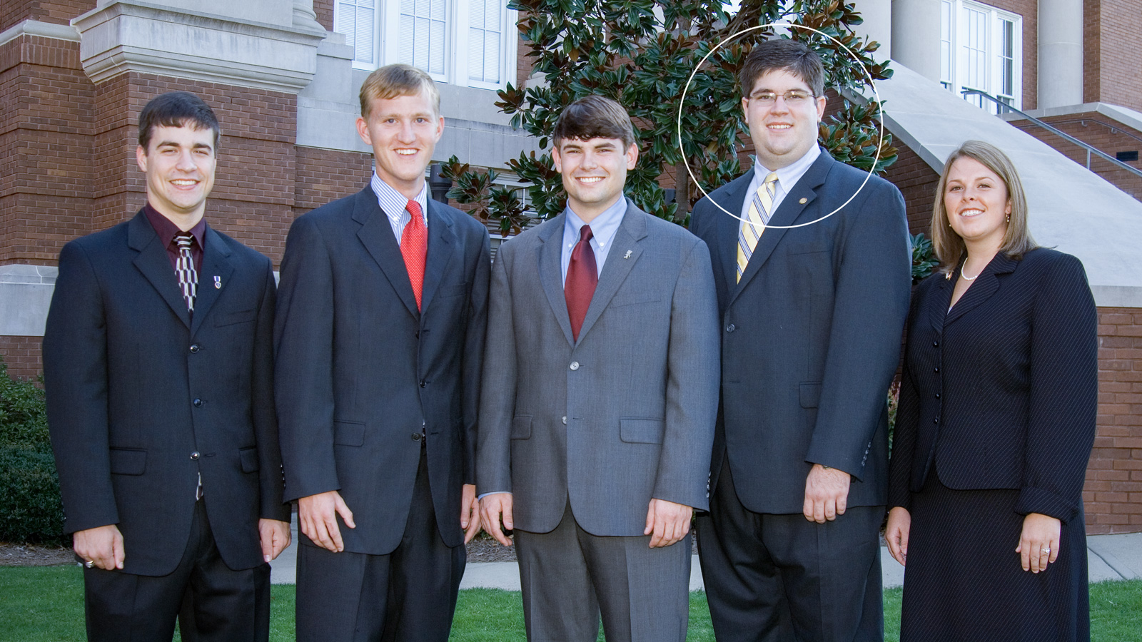 Five people in business attire stand in front of a brick building; the fourth person from the left is highlighted with a white circle.