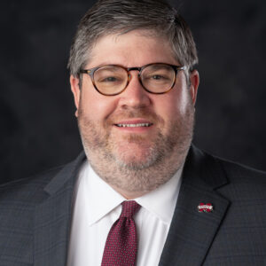 A man with short gray hair, glasses, and a trimmed beard wearing a dark suit, white shirt, and red tie, posing in front of a dark background.