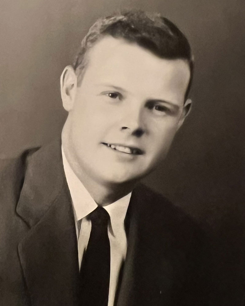 Black-and-white portrait of a young man wearing a suit and tie, looking slightly to the side and smiling gently at the camera.