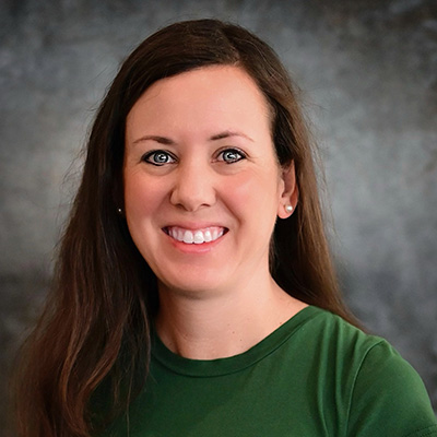 A woman with long brown hair wearing a green top smiles in front of a mottled gray background.