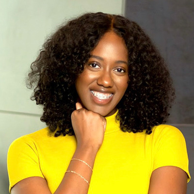 Woman with curly hair wearing a bright yellow top smiles at the camera, resting her chin on her hand. The background is neutral and softly lit.