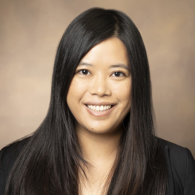 A woman with long black hair smiles at the camera against a plain, beige background. She is wearing a black top.