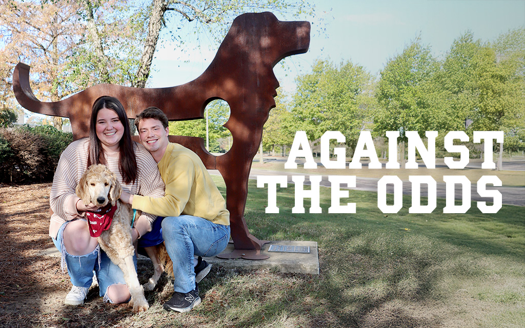 Two people kneel next to a dog wearing a bandana in front of a large metal dog statue outdoors with the text "AGAINST THE ODDS" beside them.