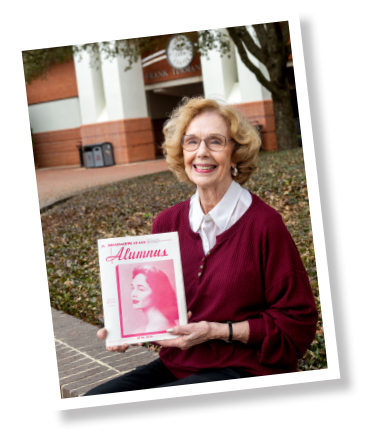 An older woman with glasses sits outdoors, smiling and holding a magazine with a vintage photo labeled "Alumnus" on the cover. A campus building is visible in the background.