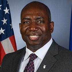 Smiling man in a suit and tie stands in front of an American flag and a blue background.