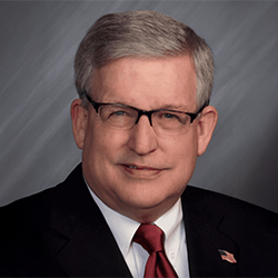 A middle-aged man with gray hair and glasses wears a dark suit, white shirt, and red tie, posing against a gray background. An American flag pin is on his lapel.