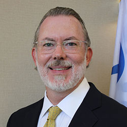 A man with gray hair, glasses, and a beard, wearing a dark suit, white shirt, and yellow tie, stands in front of a light-colored wall and a partially visible flag.