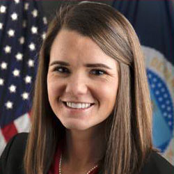 Woman with straight brown hair smiling, wearing a dark blazer and red top, standing in front of a U.S. flag and another official flag.