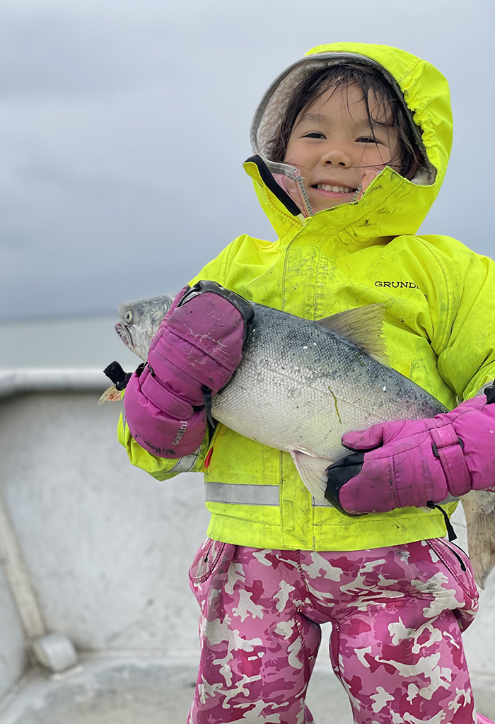 A child in a yellow rain jacket and pink gloves stands on a boat, holding a large fish and smiling at the camera.