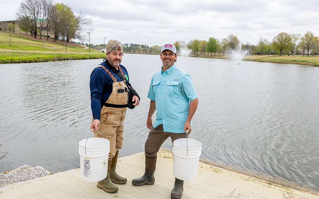 Two men in outdoor clothing and boots stand beside a lake, each holding a white bucket, with water fountains and trees visible in the background.