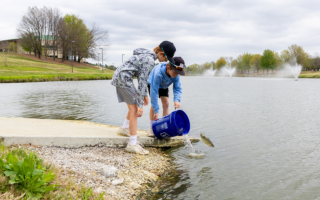 Two boys release fish from a blue bucket into a pond near a shoreline, with fountains and trees visible in the background.