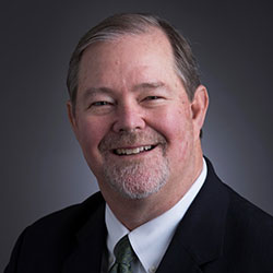 Middle-aged man with short gray hair, mustache, and goatee, wearing a dark suit, white shirt, and green tie, smiling in front of a plain dark background.