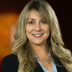 A woman with long blonde hair, wearing a black blazer and light blue blouse, smiles in front of a blurred warm-toned background.
